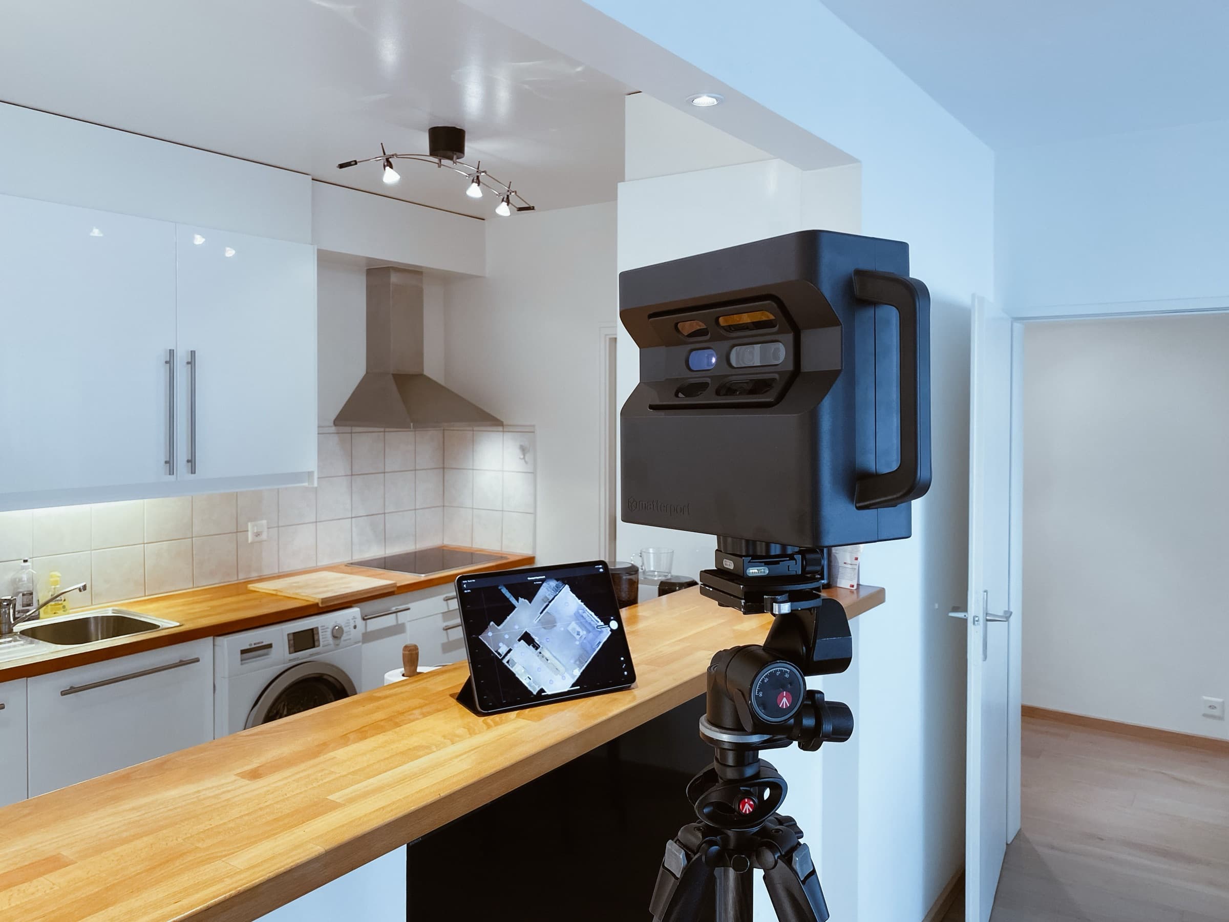 A 3D scanning camera on a tripod in a kitchen, with a tablet on the worktop showing the scanned floor plan