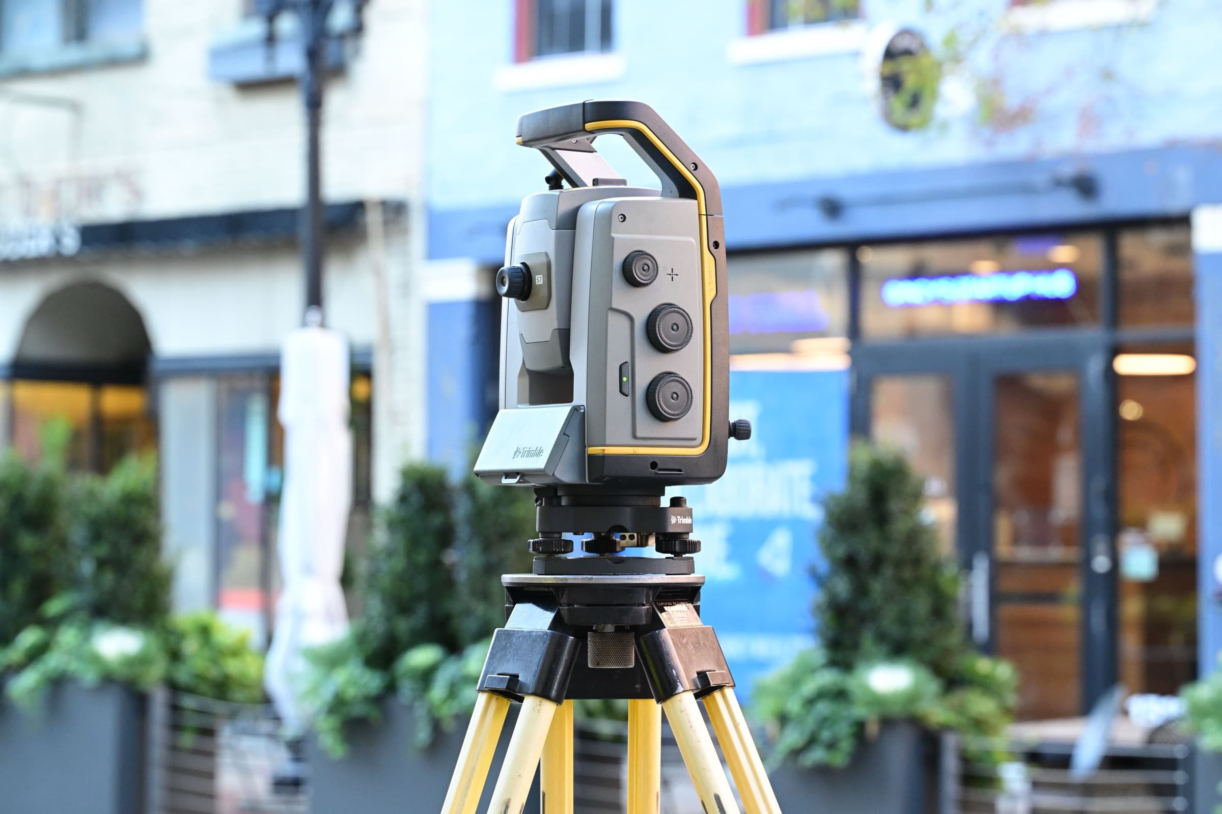 A surveying instrument on a yellow tripod set up in the street outside a row of shops